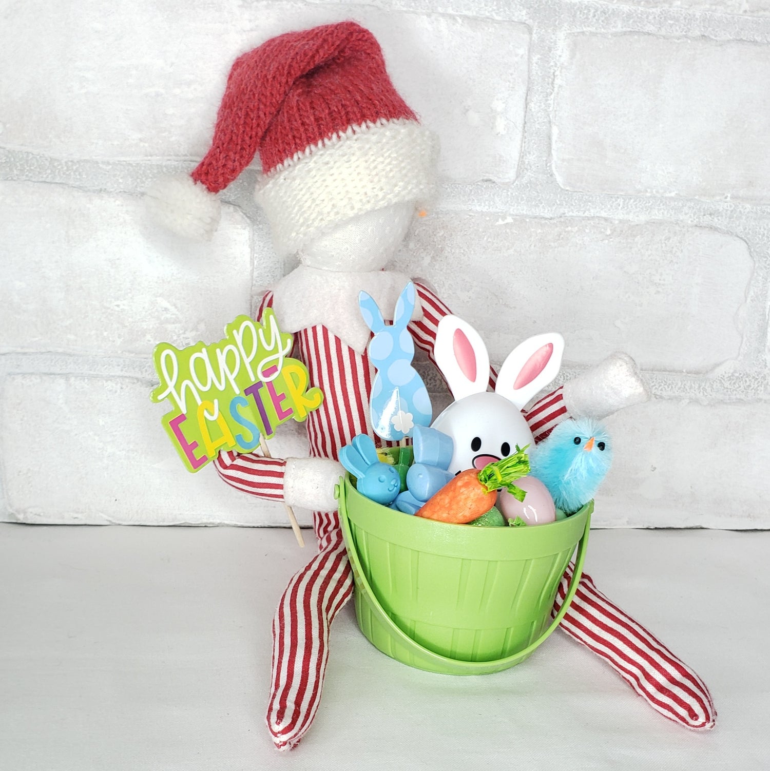 A collection of Easter-themed items including a green basket with grass, two chicks, three small Easter eggs, a bunny egg, and signs that say 'Happy Easter' and 'Bunny'. These items are arranged against a white brick background with a red and white striped fabric.