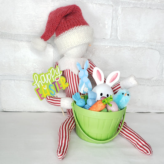 A collection of Easter-themed items including a green basket with grass, two chicks, three small Easter eggs, a bunny egg, and signs that say 'Happy Easter' and 'Bunny'. These items are arranged against a white brick background with a red and white striped fabric.
