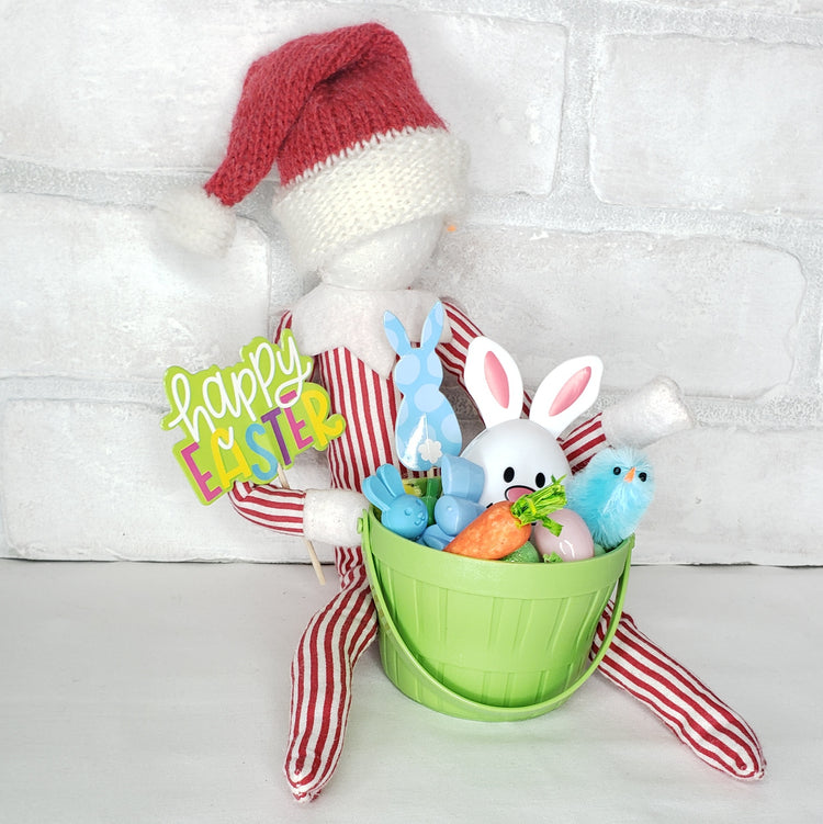A collection of Easter-themed items including a green basket with grass, two chicks, three small Easter eggs, a bunny egg, and signs that say 'Happy Easter' and 'Bunny'. These items are arranged against a white brick background with a red and white striped fabric.